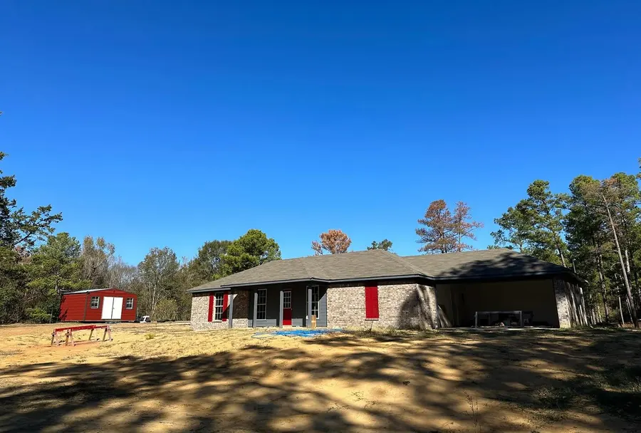 Gray brick home with red shutters and a matching red storage shed on a sunny wooded lot